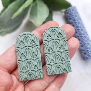 Puede incluir: Un par de pendientes de arcilla verde con un patrón geométrico. Los pendientes tienen forma de arco y una superficie texturizada.