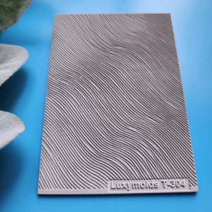 Puede incluir: Molde de silicona gris con una superficie texturizada ondulada. El molde está etiquetado como "Luxymolds T-304".