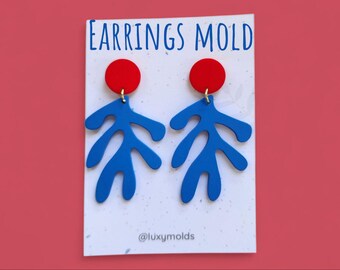 May include: A pair of blue leaf-shaped earrings with red circular tops, displayed on a white card with the text "EARRINGS MOLD" in blue. The earrings are suspended from gold-colored hooks. The background is a solid red.
