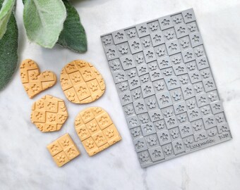Könnte beinhalten: Eine Silikonform mit einem karierten Muster und kleinen Blumendesigns. Die Form ist grau und hat den Text "Luxymolds T370" in der unteren rechten Ecke. Neben der Form befinden sich fünf kleine, gelbe, geformte Tonstücke.