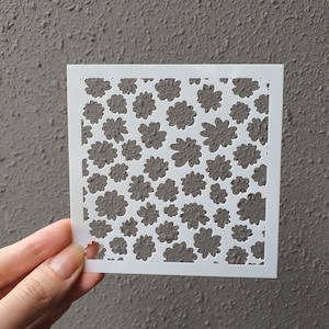 Puede incluir: Plantilla blanca con un patrón repetitivo de pequeñas formas de flores. La plantilla está hecha de un material delgado y flexible y está diseñada para usarse con pintura u otros medios.