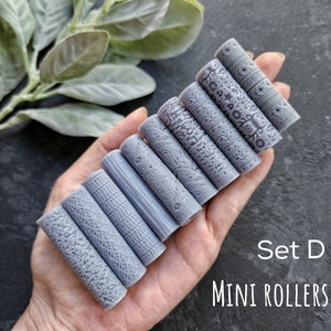 Op de afbeelding: Set van acht grijze mini-rollers met verschillende patronen. De rollers zijn in een hand gerangschikt, met de tekst "Set D MINI ROLLERS" eronder.