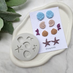 Puede incluir: Un molde de silicona para hacer joyas de resina con una variedad de formas con temática marina, incluyendo conchas marinas, estrellas de mar y un dólar de arena. El molde es gris y tiene una superficie lisa. La imagen también muestra varias piezas de resina terminadas en varios colores, incluyendo azul, marrón y morado.