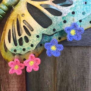 May include: Three pairs of crocheted flower earrings. One pair is pink, one pair is blue, and one pair is yellow. The earrings are hanging from a metal butterfly cutout.