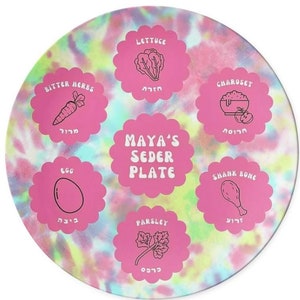 May include: A pink and blue tie-dye Passover Seder plate with six sections. Each section features a different symbol of the Seder, including lettuce, bitter herbs, charoset, shank bone, egg, and parsley. The plate is labeled "MAYA'S SEDER PLATE".
