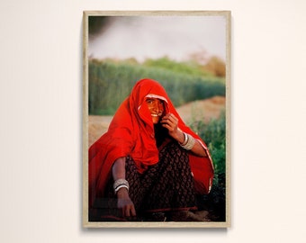 Puede incluir: Fotografía enmarcada de una persona con un velo rojo y ropa estampada, sonriendo. La persona está adornada con pulseras de plata. El fondo muestra un campo de plantas verdes. La fotografía se muestra en una pared blanca sobre una mesa marrón oscuro con tres jarrones azules.