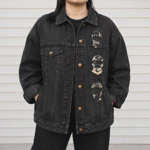 以下が含まれることがあります： クラシックな襟、長袖、フロントに金属製のボタンが付いた黒のデニムジャケット。左胸には、バラクラバマスクの模様が描かれた3つの縦型パッチが装飾として施されています。