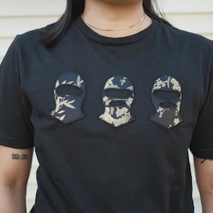 May include: A black short-sleeved t-shirt featuring three embroidered patches across the chest. Each patch is shaped like a balaclava mask and displays a black fabric base with a gold-colored floral and botanical pattern printed on the surface.