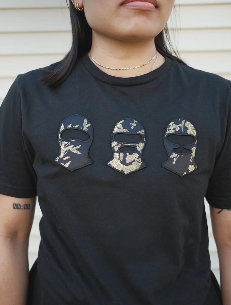 以下が含まれることがあります： 胸元に3つの刺繍パッチがあしらわれた黒の半袖Tシャツ。各パッチは目出し帽の形をしており、黒い生地をベースに金色の花や植物の模様が表面にプリントされています。