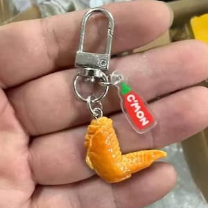 May include: A silver-toned metal keyring featuring a realistic orange-coloured chicken wing charm and a small, clear acrylic tag shaped like a hot sauce bottle with a green cap and the red text C'MON printed on the front of the bottle charm.