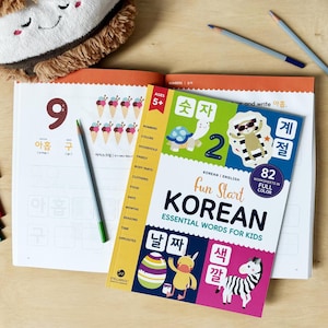Peut inclure: Un livre ouvert avec le chiffre 9 et des caractères coréens, un cahier d'exercices "Fun Start Korean" et des crayons de couleur. La couverture du cahier présente une tortue, un zèbre et le texte "Essential Words for Kids."
