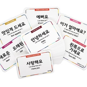 May include: A set of colorful flashcards with Korean words and their English translations. The cards feature various categories, including food and dining, everyday life, verbs and adjectives, shopping, and travel and directions. The cards are designed to help learners of the Korean language.