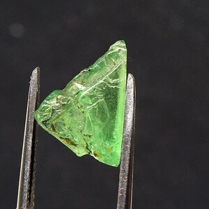 May include: A raw, translucent, light green gemstone fragment with an irregular, angular shape and a faceted, crystalline texture. The stone is held between the two metal tips of a pair of tweezers, showcasing its vibrant colour and clarity.