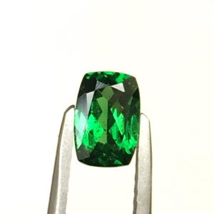 May include: A cushion-cut gemstone with a vibrant, deep green color and a faceted surface that reflects light. The stone is held by a pair of silver-toned metal tweezers, showcasing its transparent quality and precise geometric cut.