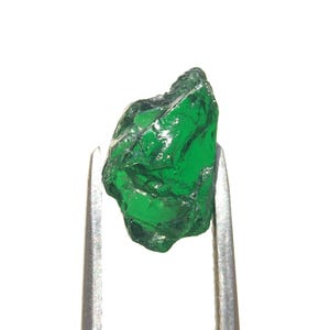 May include: A raw, uncut emerald gemstone with a deep, vibrant green colour and a translucent, crystalline structure. The stone has an irregular, jagged shape with multiple facets and natural edges, held securely by silver-toned metal tweezers.