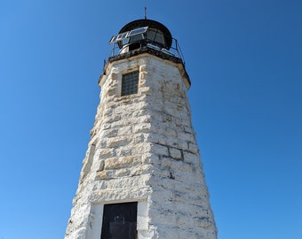 Può includere: Un alto faro in pietra bianca con una porta nera e una finestra di vetro. Il faro ha una parte superiore nera con pannelli di vetro e una piccola antenna. Il cielo è azzurro.