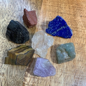 Peut inclure: Une collection de sept pierres roulées de différentes couleurs, dont le noir, le brun, le rouge, le blanc, le bleu, le vert et le violet. Les pierres sont disposées en cercle sur une surface en bois.