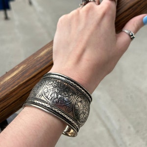 May include: A silver cuff bracelet with a decorative design of repeating circles. The bracelet is worn on a person's wrist.
