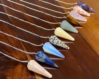 May include: A selection of gemstone pendulums with silver chains, displayed on a dark wooden surface. Each pendulum has a distinct colour and pattern. Gemstones include amethyst, rose quartz, and lapis lazuli. The pendulums are ideal for divination, each with a unique design.