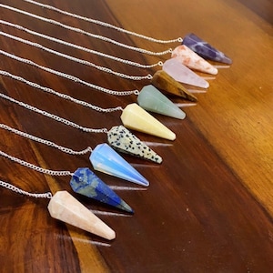 Puede incluir: Un conjunto de diez péndulos de cristal con cadenas plateadas. Los péndulos están hechos de varios cristales, incluyendo amatista, cuarzo rosa, ojo de tigre, aventurina verde, citrino, jaspe dálmata, ópalo, lapislázuli y cornalina. Los péndulos están dispuestos en una fila sobre una superficie de madera.