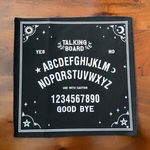 Puede incluir: Tablero Ouija negro con letras blancas y borde decorativo. El tablero presenta el texto "TALKING BOARD", el alfabeto, los números y las palabras "YES", "NO", "USE WITH CAUTION" y "GOOD BYE".