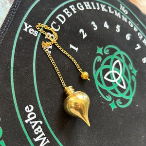Könnte beinhalten: Ein goldenes Pendel mit einer spitzen Spitze und einer Kette hängt über einem schwarzen und grünen Ouija-Brett mit den Buchstaben des Alphabets, den Zahlen 1-6 und den Wörtern "Ja", "Nein", "Vielleicht" und "Umschreiben" darauf.