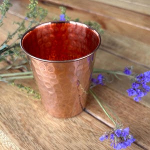 May include: A hammered copper cup with a shiny, reddish-brown finish. The cup is sitting on a wooden surface with purple flowers.