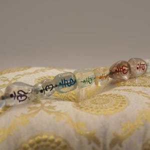 Pode incluir: Um fio de sete contas de vidro transparente com marcas de cores diferentes. Cada conta tem um símbolo sânscrito inscrito nela. As contas estão apoiadas em um tecido branco com um padrão dourado.
