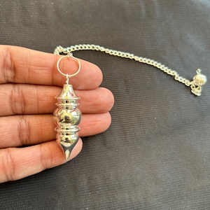 Könnte beinhalten: Ein silbernes Metallpendel mit einer spitzen Spitze und einer Kette. Das Pendel hat drei abgerundete Abschnitte.