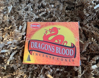 Może przedstawiać: Pudełko stożków kadzidła "Dragons Blood". Pudełko jest pomarańczowe i czerwone z żółtym wzorem słońca i ilustracją czerwonego smoka. Pudełko otoczone jest brązowym wypełniaczem papierowym.