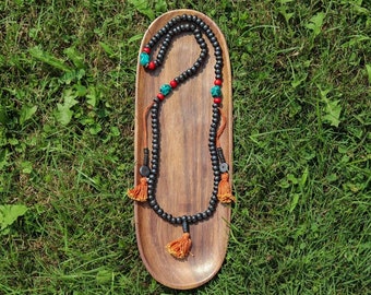 May include: A black wooden mala bead necklace with turquoise and red accents. The necklace is displayed on a wooden tray with orange tassels.