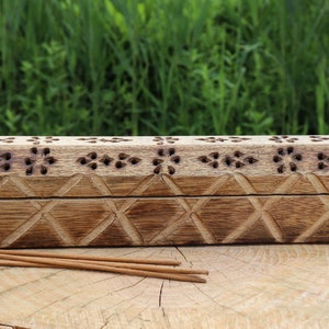 May include: A wooden incense holder with intricate carvings. The holder has a rectangular shape with a decorative pattern of diamonds and flowers. It is made of natural wood and has a rustic brown colour.