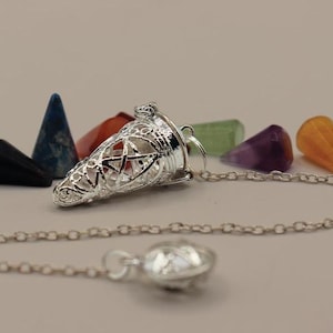 May include: A silver pendulum with a delicate, intricate design and a chain. The pendulum is shaped like a cone and has a star-shaped design at the top. The chain is attached to a small, round, silver weight. The pendulum is surrounded by seven small, colorful crystals.