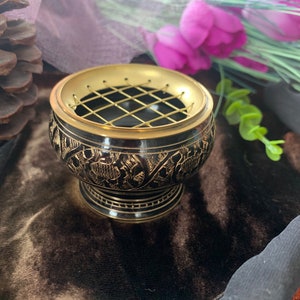 Puede incluir: Un quemador de incienso de metal negro y dorado con un intrincado diseño floral. El quemador tiene una rejilla de alambre para quemar conos de incienso.