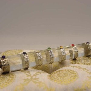Pode incluir: Uma varinha de cristal de quartzo transparente com faixas prateadas e pedras preciosas coloridas. A varinha está apoiada em um tecido branco e dourado com padrões.