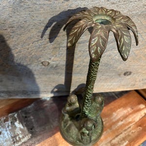 May include: A bronze-colored palm tree candle holder. The palm fronds form the top, with a textured trunk and a base featuring detailed monkey figures. The candle holder is made of metal and has a vintage appearance.