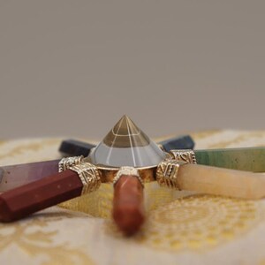May include: A clear crystal pyramid with a gold base sits in the centre of a seven-pointed star made of polished gemstones. The gemstones are amethyst, red jasper, carnelian, citrine, green aventurine, blue sodalite, and clear quartz.