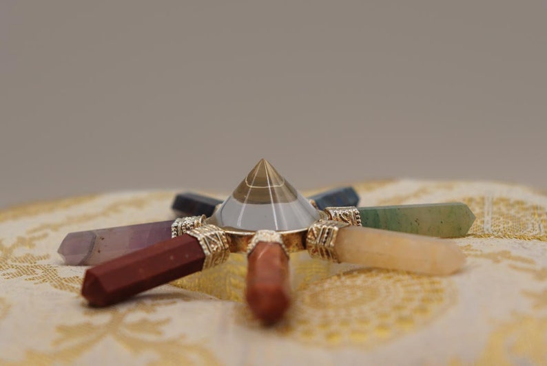 May include: A clear crystal pyramid with a gold base sits in the centre of a seven-pointed star made of polished gemstones. The gemstones are amethyst, red jasper, carnelian, citrine, green aventurine, blue sodalite, and clear quartz.