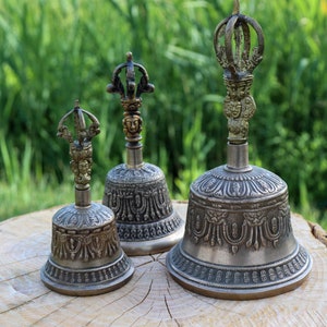 May include: Three antique silver-colored bells with ornate detailing and decorative handles. The bells are of varying sizes, with intricate patterns on the bell's body and a detailed crown-like top. The bells are displayed on a wooden surface against a green background.
