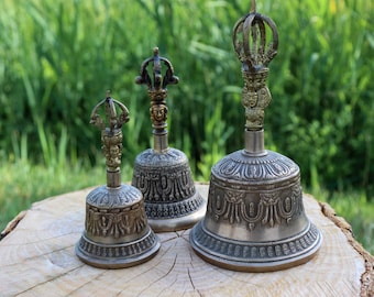 May include: Three antique silver-colored bells with ornate detailing and decorative handles. The bells are of varying sizes, with intricate patterns on the bell's body and a detailed crown-like top. The bells are displayed on a wooden surface against a green background.