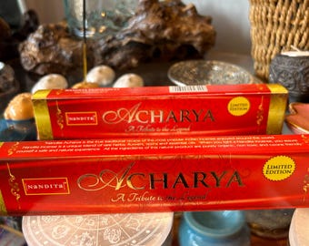Puede incluir: Dos cajas de inciensos en forma de palo con un diseño rojo y dorado. Las cajas están etiquetadas con "Acharya" y "A Tribute to the Legend". Las cajas están sobre una mesa con una superficie de vidrio transparente.