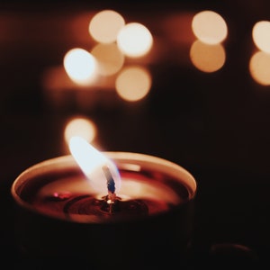 May include: A single candle with a red wax base and a blue wick is lit and burning. The candle is in focus, while the background is out of focus and shows a warm glow of other candles.