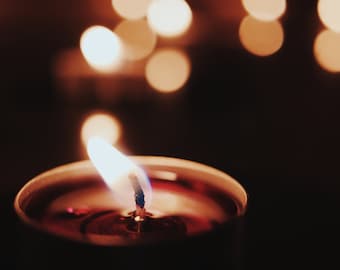 May include: A single candle with a red wax base and a blue wick is lit and burning. The candle is in focus, while the background is out of focus and shows a warm glow of other candles.