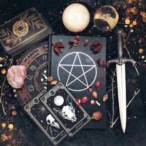 Puede incluir: Un libro negro con un símbolo de pentagrama y pétalos de rosa roja en la portada. También hay un puñal de plata, una bola de cristal, una esfera blanca y una baraja de tarot en la imagen.