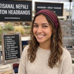 Op de afbeelding: Een vrouw draagt een bordeauxrode katoenen hoofdband en glimlacht. Het bord achter haar zegt "ARTISANAL NEPALI COTTON HEADBANDS". Een kleiner bord geeft de beschikbare kleuren weer: Kobaltblauw, Haver, Terracotta, Heather Grey, Zuiver Wit, Donkere Chocolade, Petrolgroen, Jet Chocolatk, Diep Bourgondië, Olijfgroen en Schuimblauw.