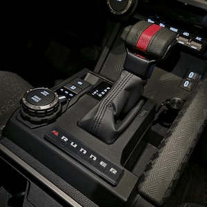 May include: Interior view of a vehicle's console, featuring a black leather gear shift with a red stripe. The console includes a "RUNNER" emblem, drive mode controls, and USB ports. The overall design is modern and sleek.