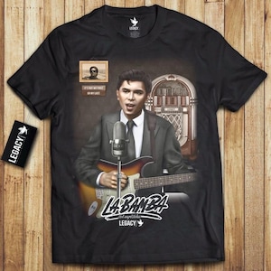 May include: Black t-shirt featuring a graphic of a singer playing a guitar, with a vintage microphone and a jukebox in the background. The text "La Bamba" and "Legacy" are also visible.