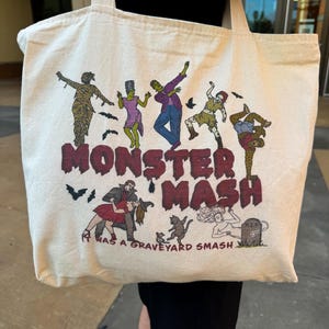 May include: A white canvas tote bag with a colorful illustration of monsters dancing. The text "Monster Mash" is printed in red and black. The text "It was a graveyard smash" is printed below the illustration.