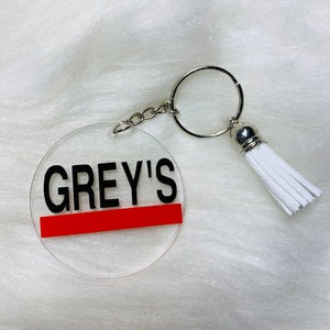 May include: A clear acrylic keychain with a red stripe and the text "GREY'S" in black lettering. The keychain has a silver metal ring and a white tassel.