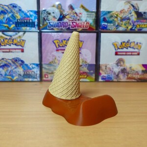 May include: A brown and tan ice cream cone holder. The holder has a brown base with a textured design, and a tan cone-shaped top. The holder is designed to hold an ice cream cone. The background features boxes of trading cards.